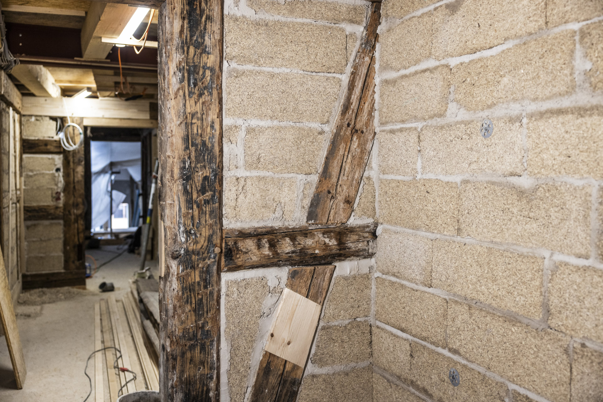 Detailansicht einer Fachwerkwand Detailansicht einer Lehmziegel-FachwerkwandDetailaufnahme einer sanierten Fachwerkwand mit Lehmziegeln und freigelegtem Holzgebälk in einem Innenraum, sichtbare Baustellenmaterialien im Hintergrund.